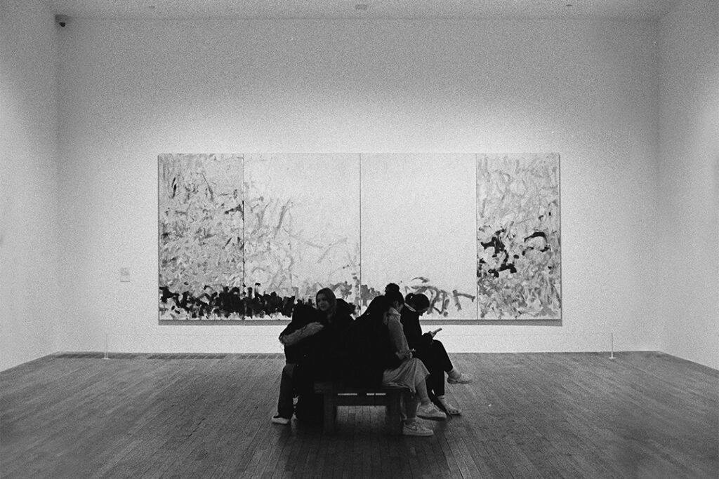 A black and white photo of a group of students sitting in front of a large abstract painting.