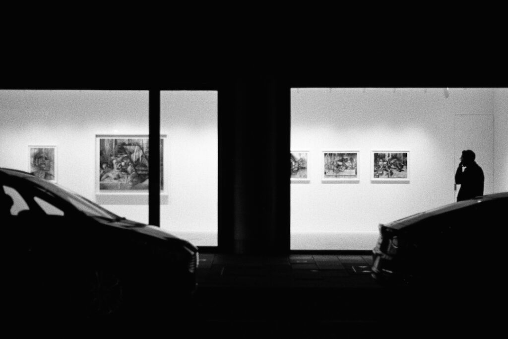 A black and white photo of a modern art gallery at night. Its lights shine bright and put passers-by in strong silluette.