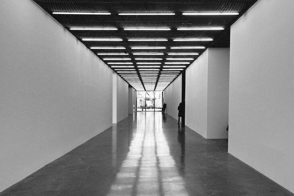 The empty interior corridor of a contemporary art gallery. The strong geometric shapes of the gallery space dwarf the figures within.