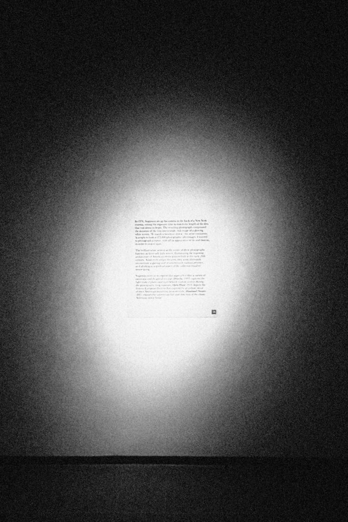 An black and white poster of the accompanying text to an art exhibition, illuminated by a strong spotlight. The text is illegible.