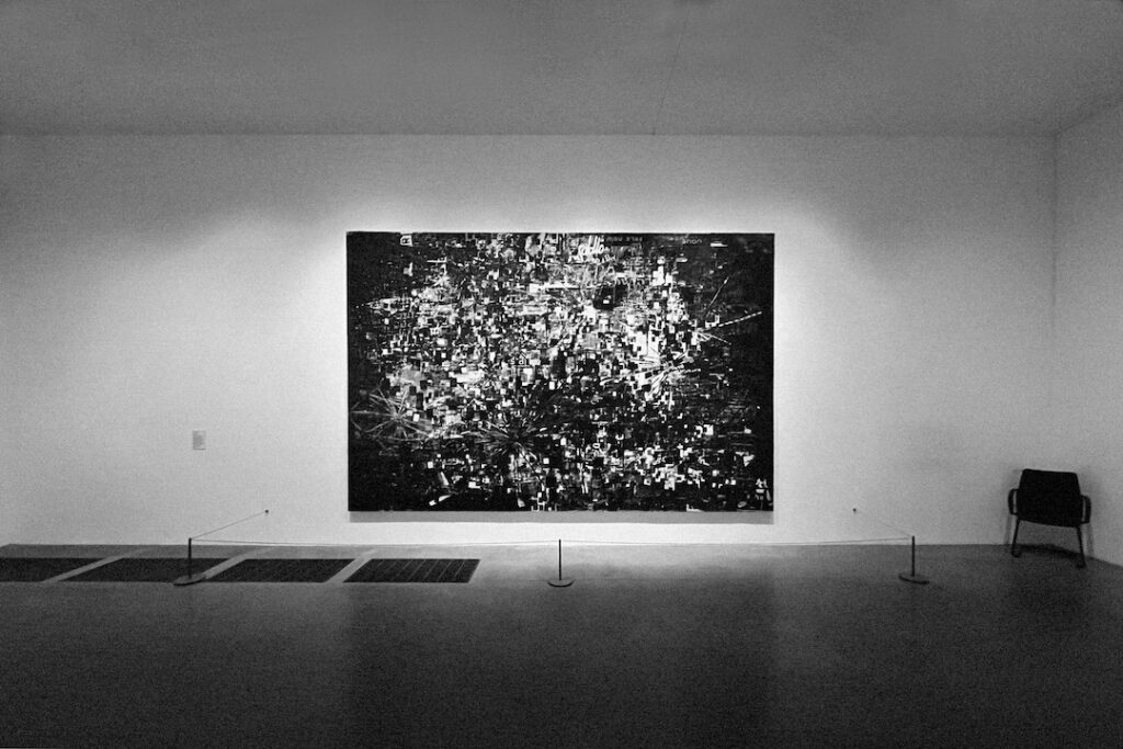 A black and white photo of a large painting hanging in an art gallery with an empty invigulator's chair to the side.