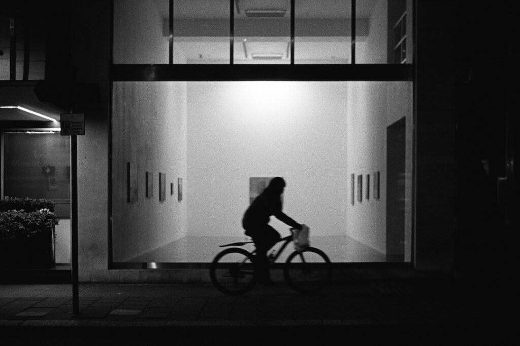 A black and white photo of a cyclist passing a modern art gallery at night. The bright lights of the gallery illuminate the scene and cast the cylist into shadow.